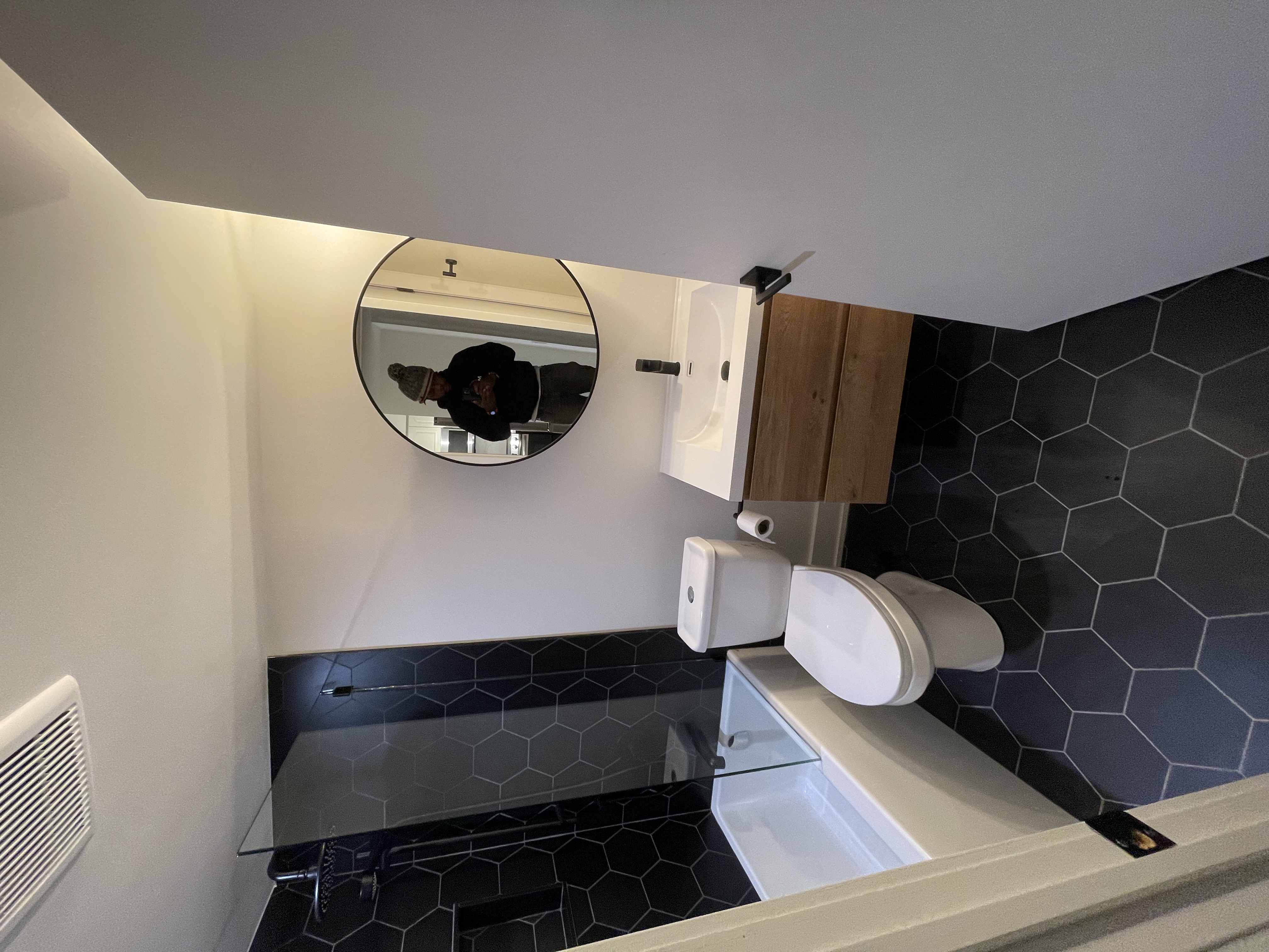 Modern bathroom with striking black hexagon tile and warm wood vanity — strong portfolio piece despite photographer reflection — Queensway, Hamilton | MTC Renovations